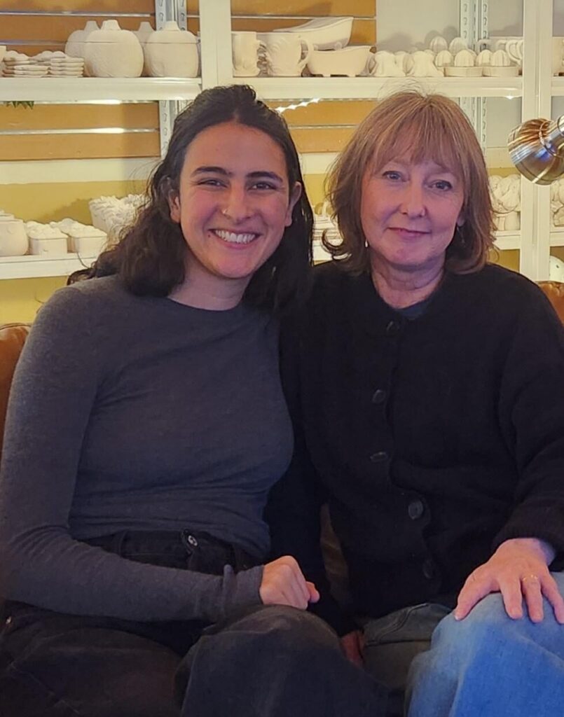 Two women sit next two each other and look at the camera with their legs crossed
