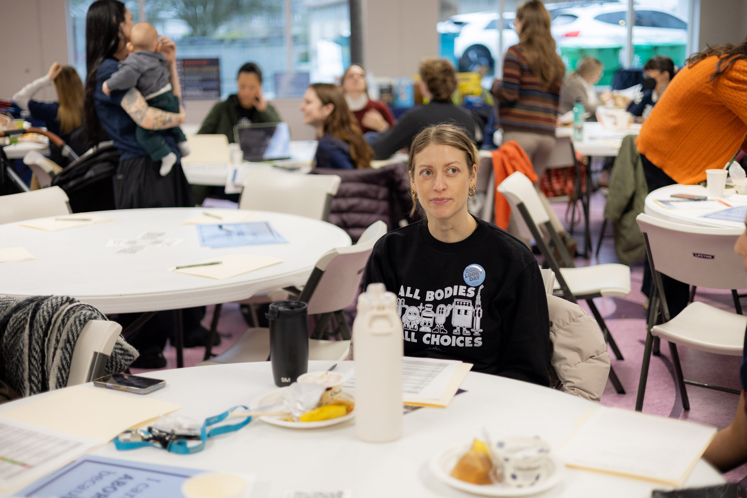 A room of with tables and chairs with one women in the center looking off to the side. Her shirt says All Bodies All Choices